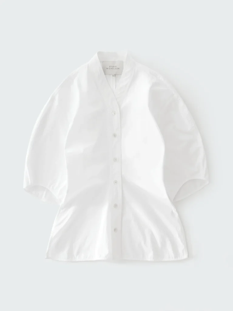 Minimalist white Studio Nicholson cotton shirt with sculptural short sleeves, elongated relaxed silhouette, soft V-neck collar, and clean button-front construction photographed against a pale gray background