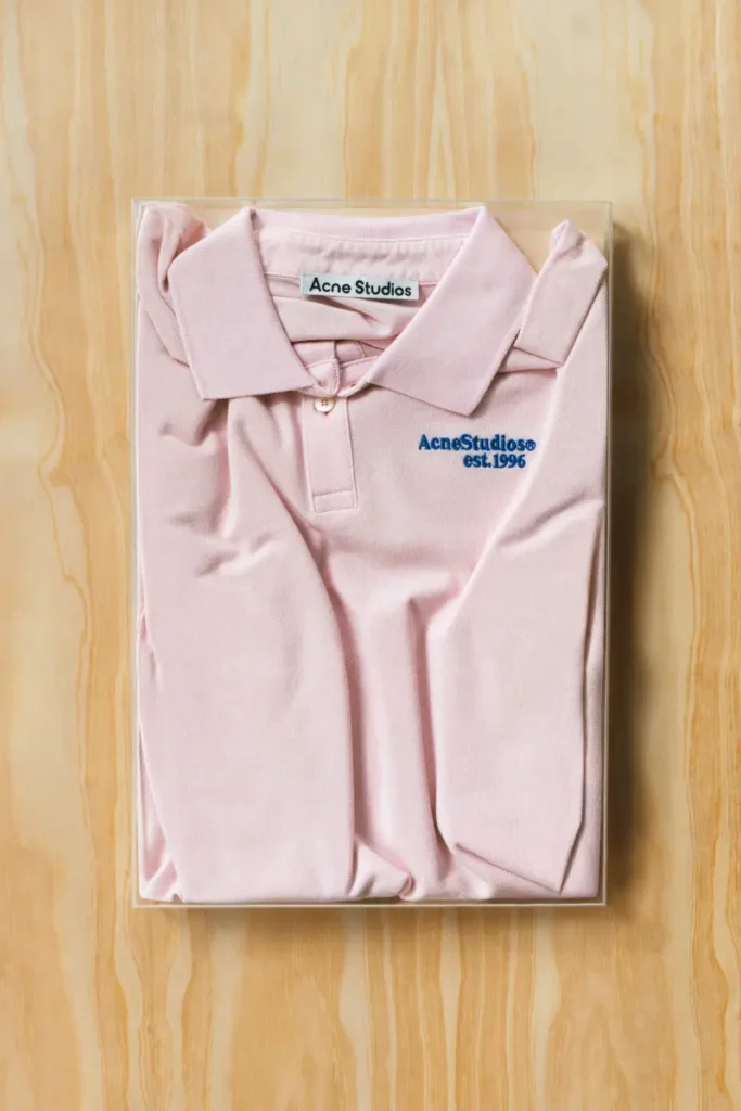 A neatly folded pale pink polo shirt is encased within a clear, box-like frame, presented against a warm wood backdrop that emphasizes its soft tonal palette. The fabric gathers gently at the folds, creating subtle shadows that lend depth to the otherwise minimal composition. At the chest, a small embroidered mark—“Acne Studios est. 1996”—adds a quiet note of identity, while the collar and placket remain crisply defined. The transparent casing elevates the garment into an object of display, where everyday wear is reframed as a considered artifact within the visual language of Acne Studios