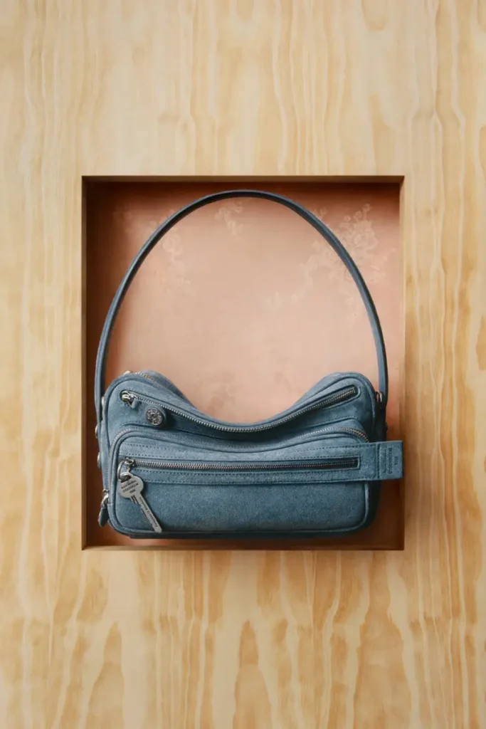 A compact blue leather shoulder bag is set within a recessed wooden frame, its curved silhouette echoing the soft arc of the strap above. The surface carries a gently worn, almost velvety texture, punctuated by multiple zip compartments and silver hardware that introduce subtle utility. Behind it, a peach-toned fabric lining adds warmth and contrast, softening the industrial precision of the bag’s construction. Suspended in this boxed composition, the accessory reads as both functional object and studied form—an understated balance of practicality and design, characteristic of Acne Studios