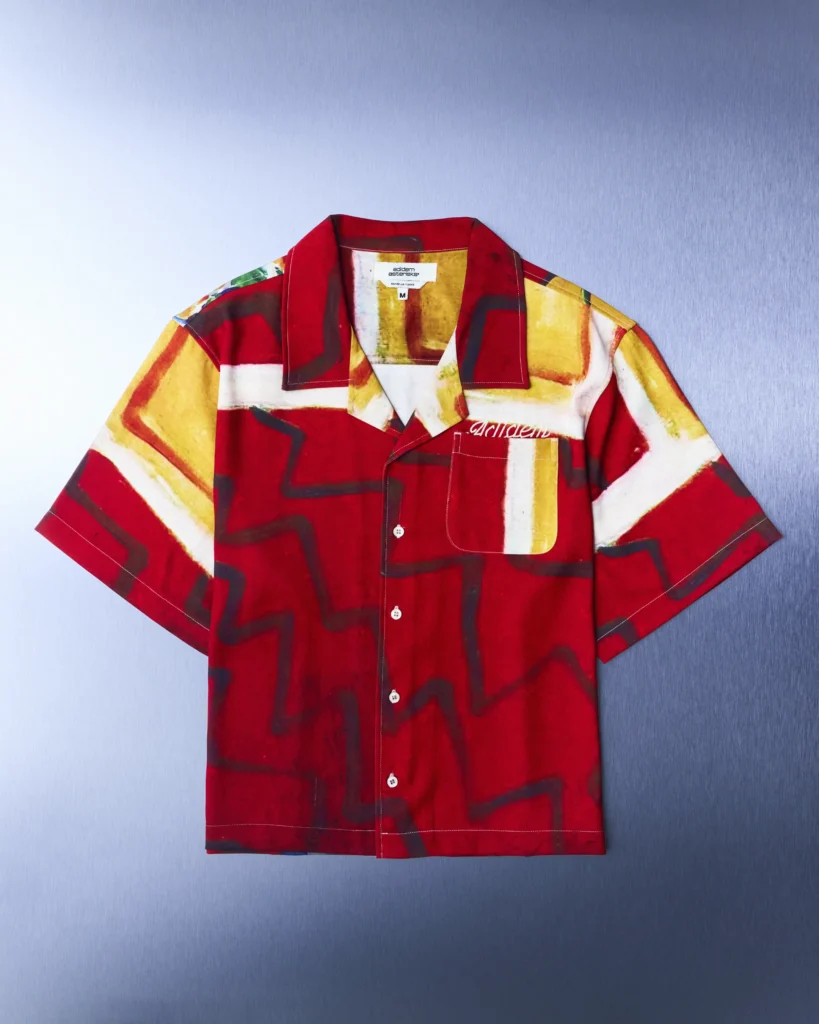 Studio product image of the Adidem Astericks Spring/Summer 2026 print camp collar shirt in a rich red colorway featuring painterly geometric blocks in yellow, cream, and charcoal tones, finished with tonal embroidered chest script, patch pocket detailing, white buttons, and a relaxed boxy short-sleeve silhouette against a metallic blue backdrop