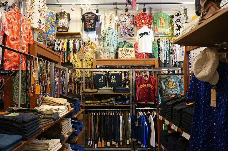 Interior of a boutique clothing store filled with neatly arranged racks and shelves of apparel. The space features a dense display of colorful patterned shirts—many with floral, graphic, and retro prints—hung along the walls and above eye level. Folded denim and trousers are stacked on wooden shelves in the foreground, while jackets, T-shirts, and accessories hang on central racks. The overall atmosphere feels vibrant and curated, with a mix of vintage-inspired and streetwear styles showcased in a compact, well-organized layout