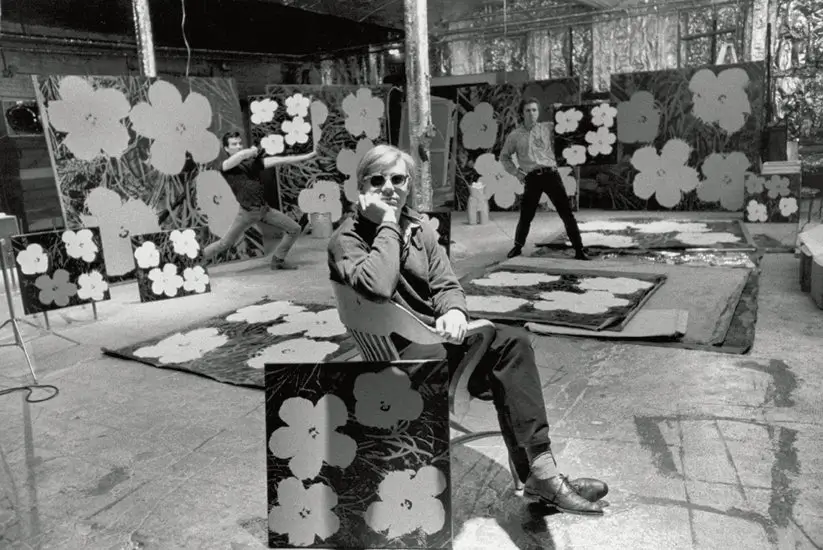 A wide black-and-white documentary-style photograph depicting a cavernous industrial studio interior transformed into an improvised creative environment. Metallic foil wraps portions of the ceiling, columns, and walls, reflecting harsh overhead light across the unfinished concrete floor and scattered equipment. In the middle of the space, a lone seated figure occupies a curved couch, bent over papers or a notebook in quiet concentration, while cables snake across the ground toward projectors, carts, and fragmented workstations positioned throughout the room.The image carries the stark utilitarian atmosphere associated with experimental art production spaces of the 1960s, particularly the raw aesthetic of Andy Warhol’s Factory. Rather than romanticizing artistic labor, the composition foregrounds the mechanics behind creation: exposed infrastructure, temporary partitions, electrical clutter, and the visible exhaustion of process. The reflective foil surfaces introduce a strange cinematic quality, making the studio feel simultaneously futuristic and deteriorated, like a hybrid between an underground film set, performance space, and industrial workshop.Large areas of negative space amplify the isolation of the central figure, whose small presence contrasts with the overwhelming architecture surrounding them. The photograph ultimately reads less as a portrait and more as an environmental study of artistic production itself—where experimentation, repetition, silence, and physical labor become embedded directly into the space