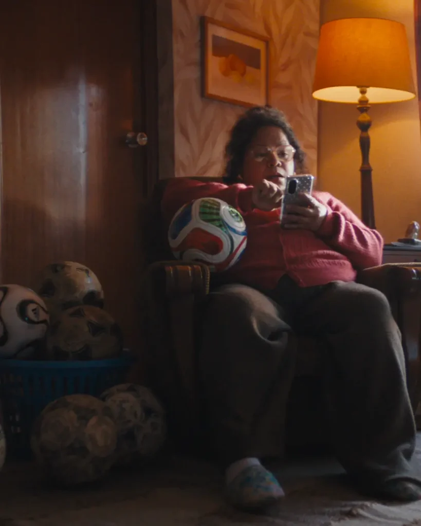 A woman sits in a dimly lit living room armchair, focused on her smartphone while surrounded by scattered footballs across the wooden floor. She wears a textured pink cardigan and dark lounge pants, illuminated by the warm glow of a tall vintage floor lamp beside her. A colorful FIFA World Cup-style football rests against the chair, while additional worn soccer balls fill a basket nearby, adding to the lived-in atmosphere. Patterned wallpaper, framed wall art, and soft cinematic shadows create a nostalgic, intimate domestic scene centered around football culture and quiet evening routine