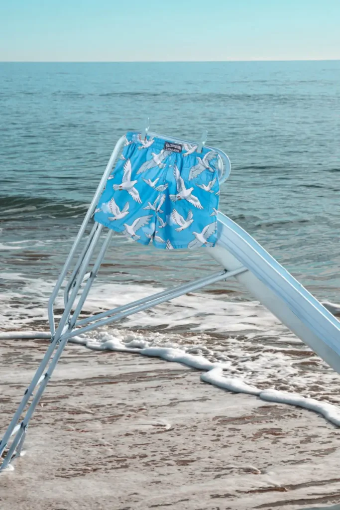 Sky-blue Vilebrequin x 3.Paradis swim shorts covered in soaring dove graphics rest against a pale seaside slide overlooking the ocean. The serene coastal setting and surreal print language evoke freedom, escapism, and the dreamlike Riviera spirit central to the Summer 2026 capsule