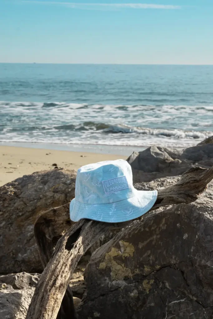 A pale blue Vilebrequin x 3.Paradis bucket hat rests on driftwood along a rocky shoreline, framed by soft ocean waves and Mediterranean light. Subtle tonal graphics and embroidered branding give the accessory a relaxed yet elevated Riviera feel, reflecting the capsule’s dreamlike vision of coastal escape and understated