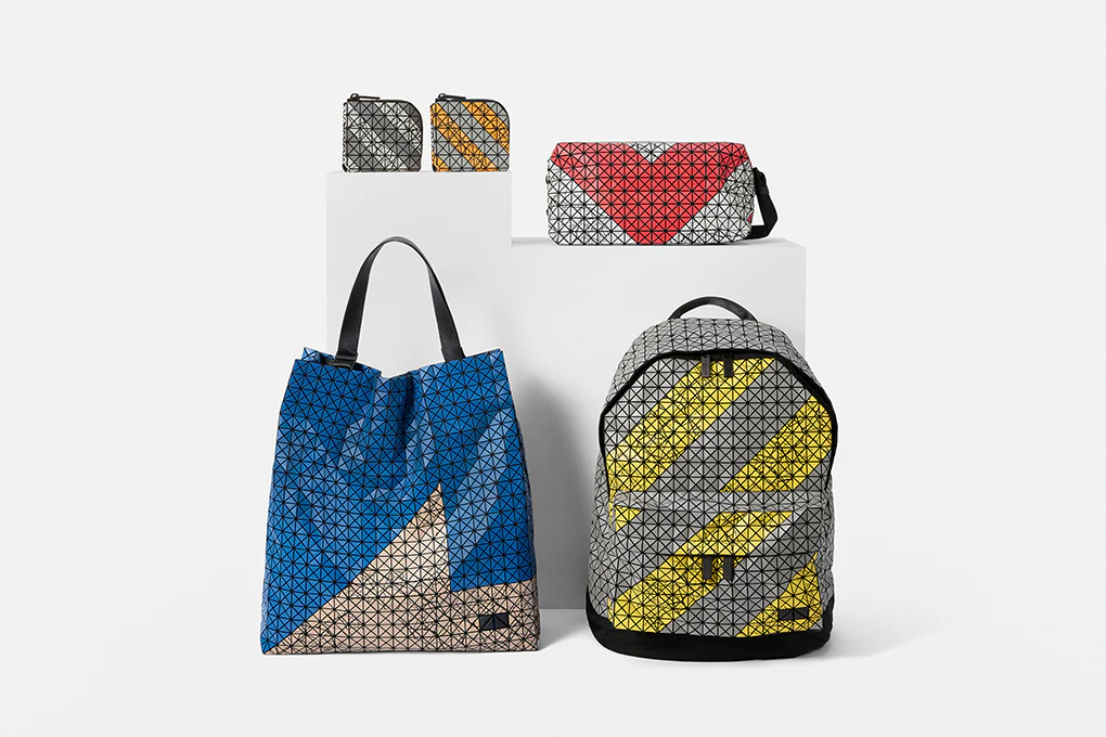 Studio display of multiple Bao Bao Issey Miyake bags from the Autobahn-inspired collection, featuring the signature triangular geometric panel construction in bold graphic colorways. A gray and yellow Daypack Autobahn backpack stands beside a blue tote, while smaller pouches and a red-and-white shoulder bag rest on elevated platforms, emphasizing the collection’s architectural, industrial-inspired aesthetic