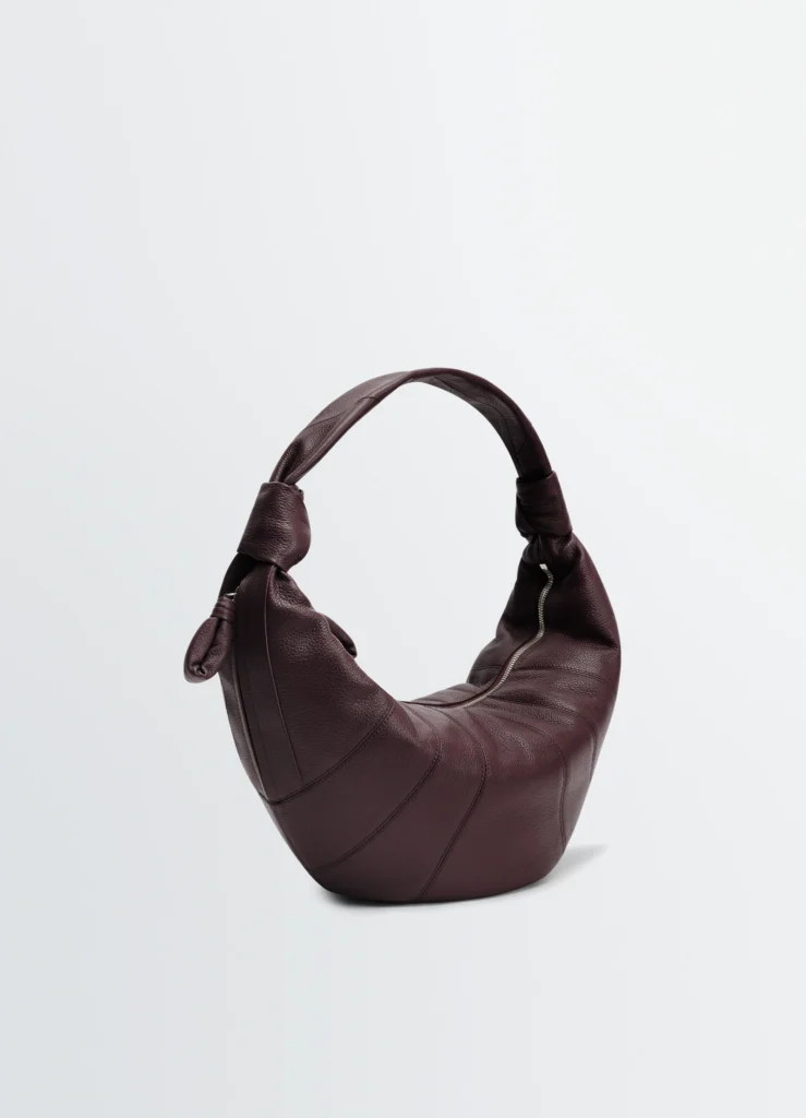 Sculptural crescent hobo bag in deep espresso-burgundy pebbled leather featuring curved paneled stitching, knotted shoulder strap details, and a visible silver zipper opening against a soft light grey studio background