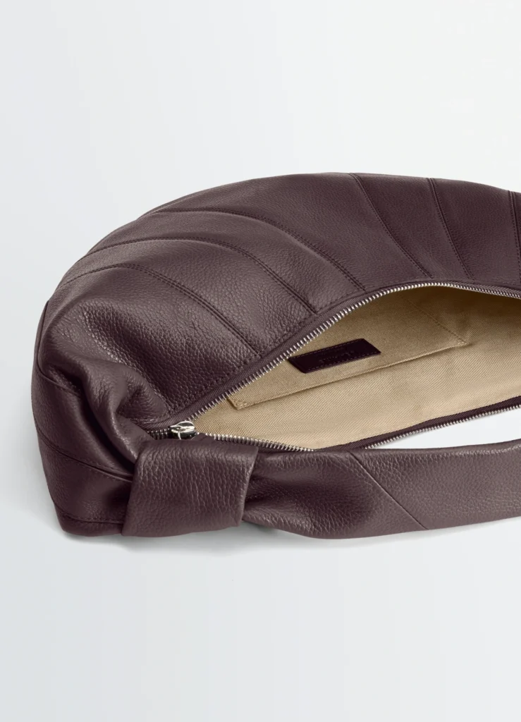 Close-up of a sculptural crescent leather shoulder bag in deep espresso-burgundy featuring curved paneled stitching, a knotted strap detail, silver zipper closure, and a beige canvas-lined interior against a clean light grey background