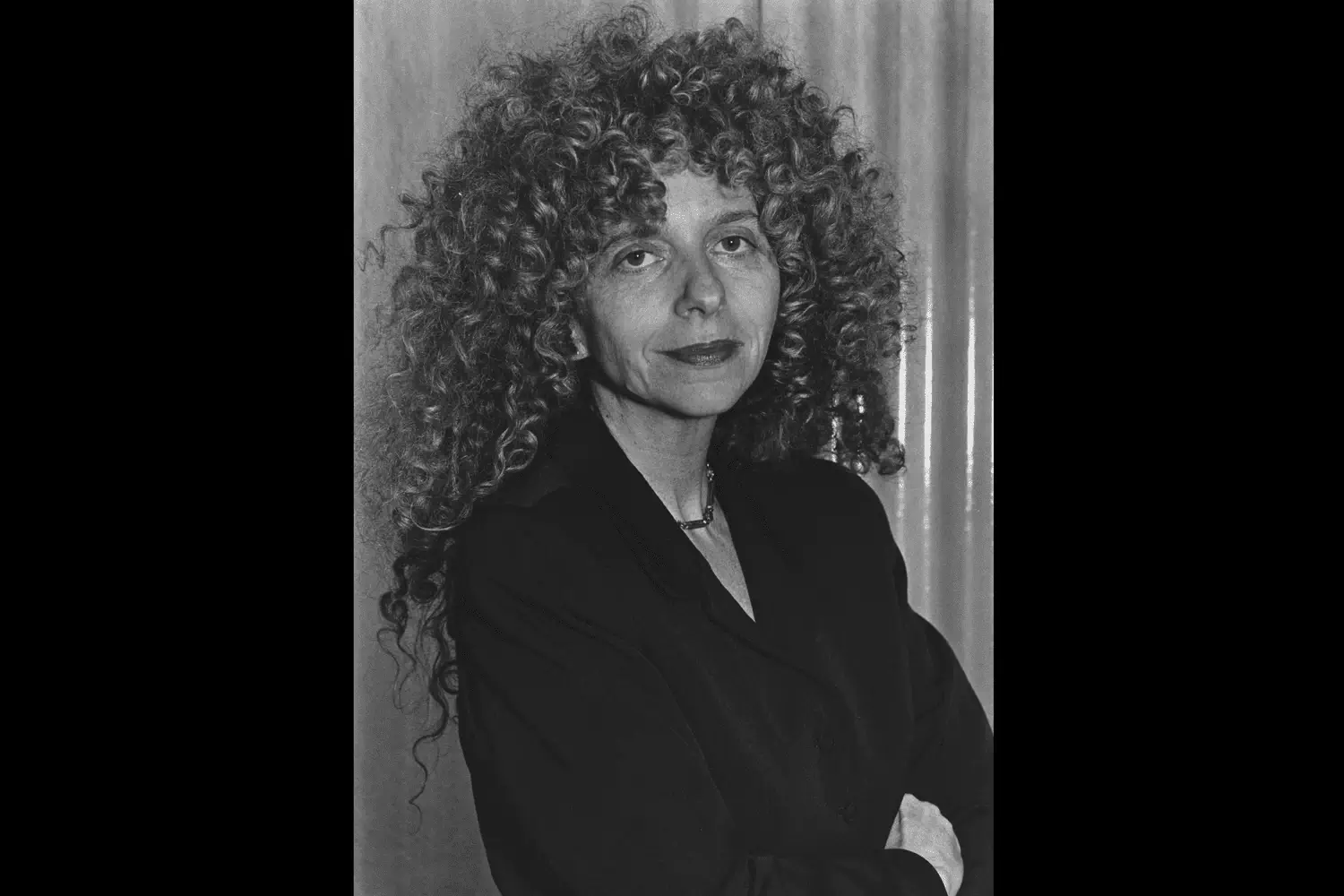 A black-and-white portrait captures Barbara Kruger with her signature voluminous curls and composed, analytical gaze, reflecting the sharp intellectual presence that reshaped feminist conceptual art in the late twentieth century. Dressed in minimalist dark tailoring, the artist appears both understated and formidable, embodying the same confrontational clarity and media-awareness that define her iconic text-based works
