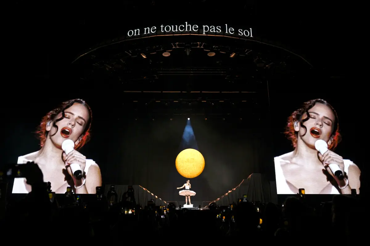 Wide arena concert scene showing a performer centered beneath a glowing oversized moon installation while twin giant screens project close-up vocals in real time. The theatrical staging combines intimate performance with monumental scale, framed by dark lighting, audience silhouettes, and the phrase “on ne touche pas le sol” suspended above the stage