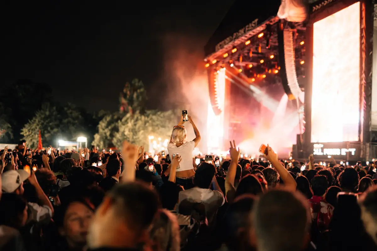Nighttime festival crowd gathered in front of a brightly illuminated main stage, with audience members raising phones to capture the performance. A concertgoer elevated above the crowd records the moment while warm orange lights, haze effects, and large LED screens create an energetic live music atmosphere