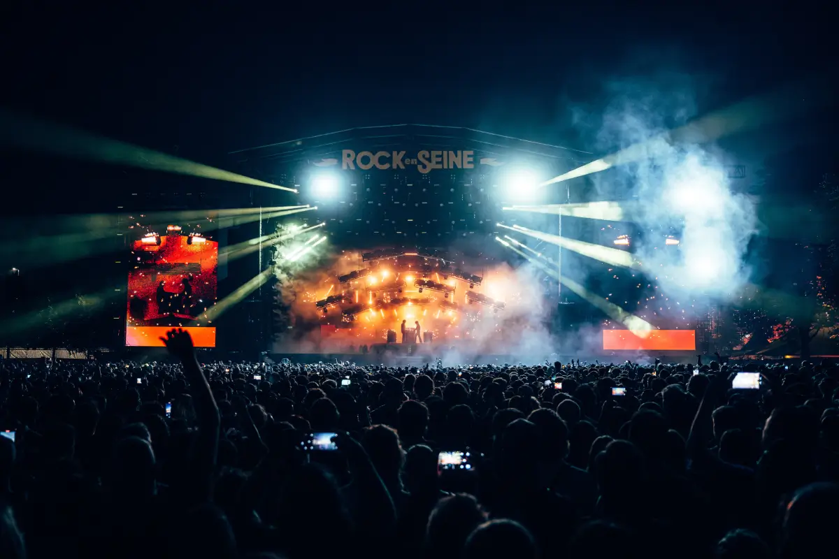 Wide-angle nighttime festival scene capturing a massive crowd facing the illuminated Rock en Seine main stage. Bright beams of light cut through smoke and haze while audience members hold phones aloft, creating an electric atmosphere of scale, anticipation, and collective live music energy