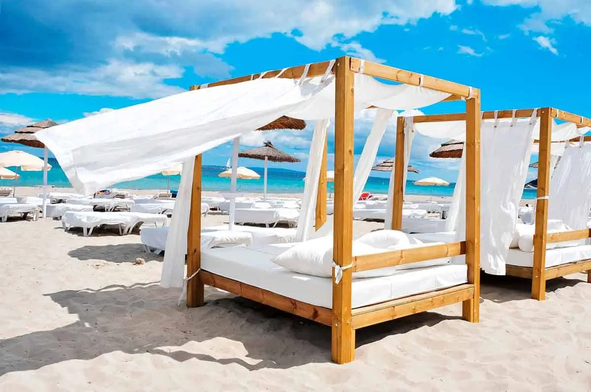 This beachside scene captures the understated luxury of contemporary Mediterranean resort culture through minimalist wooden cabanas draped in flowing white fabric. Set directly against turquoise water and bright coastal light, the space feels calm, immersive, and intentionally refined