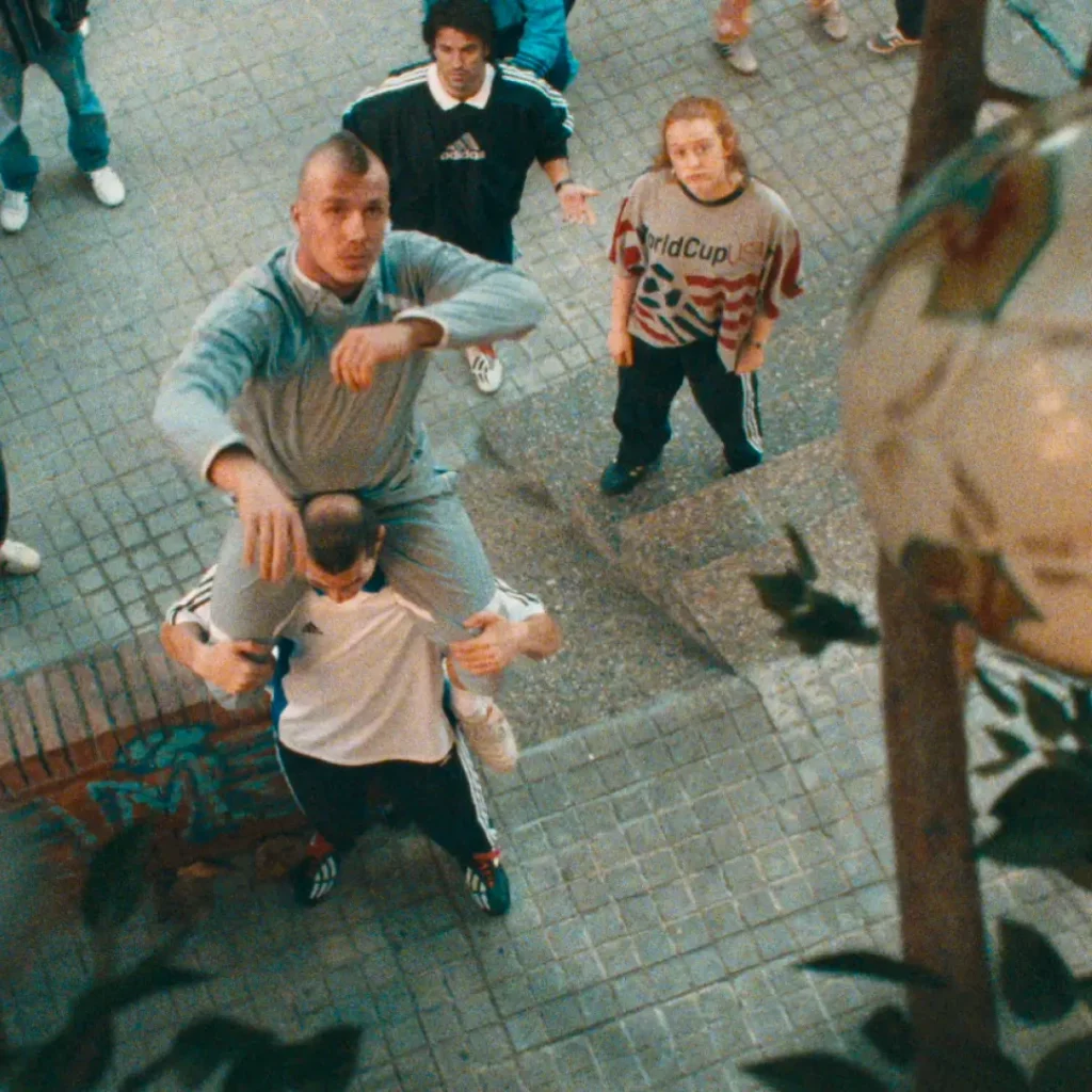 A group of young adults gather in a concrete apartment courtyard, looking upward toward something above the frame in a cinematic overhead shot. At the center, one man balances on another person’s shoulders while dressed in a light gray adidas tracksuit, surrounded by others wearing vintage-inspired football apparel and oversized sportswear. A standout graphic World Cup shirt and retro adidas polos reinforce the nostalgic street-football aesthetic. Soft grain, muted colors, and the elevated perspective create the feeling of a spontaneous neighborhood moment rooted in community, football culture, and early-2000s sports fashion