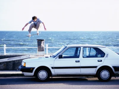 Archival skateboarding photograph showing a skater performing a high ollie above a seaside promenade obstacle while a white sedan passes in the foreground, framed against open water and bright coastal light—capturing the raw, improvisational spirit of 1990s UK street skating
