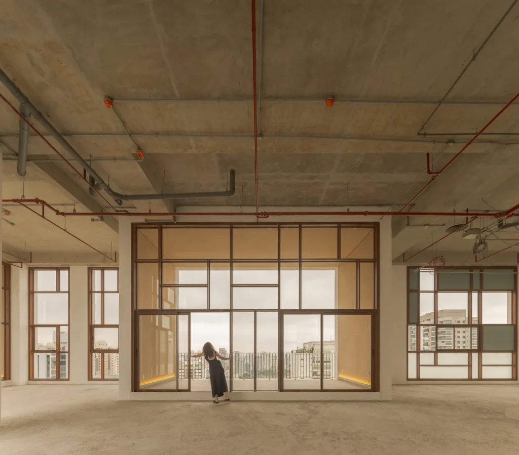 A vast unfinished loft-like interior features exposed concrete ceilings, visible industrial piping, and expansive grid-framed glass partitions surrounding a softly illuminated central living volume. A solitary figure in dark clothing stands near the floor-to-ceiling windows overlooking the city skyline, emphasizing the scale and minimalist architectural atmosphere of the raw contemporary space