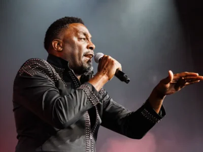 Side-profile concert portrait of a performer holding a microphone mid-performance while gesturing toward the audience. He wears a structured black stage jacket embellished with reflective studs across the shoulders, sleeves, and front placket. Warm theatrical lighting and soft haze create a dramatic atmosphere, emphasizing the expressive live-performance moment and elevated stage styling