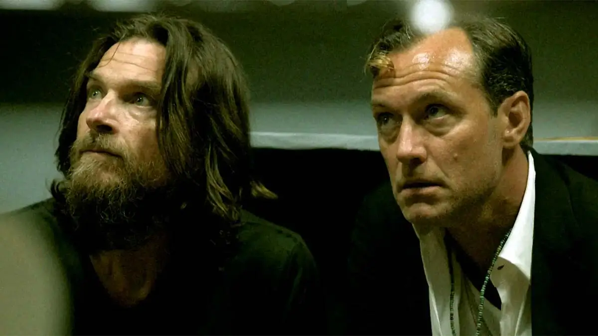 Moody cinematic still featuring two men seated side by side in a dimly lit interior, one with long hair and a beard in casual dark clothing and the other in a dark suit with an open-collar shirt. Both look upward with tense, concentrated expressions, creating an atmosphere of suspense and uneasy partnership