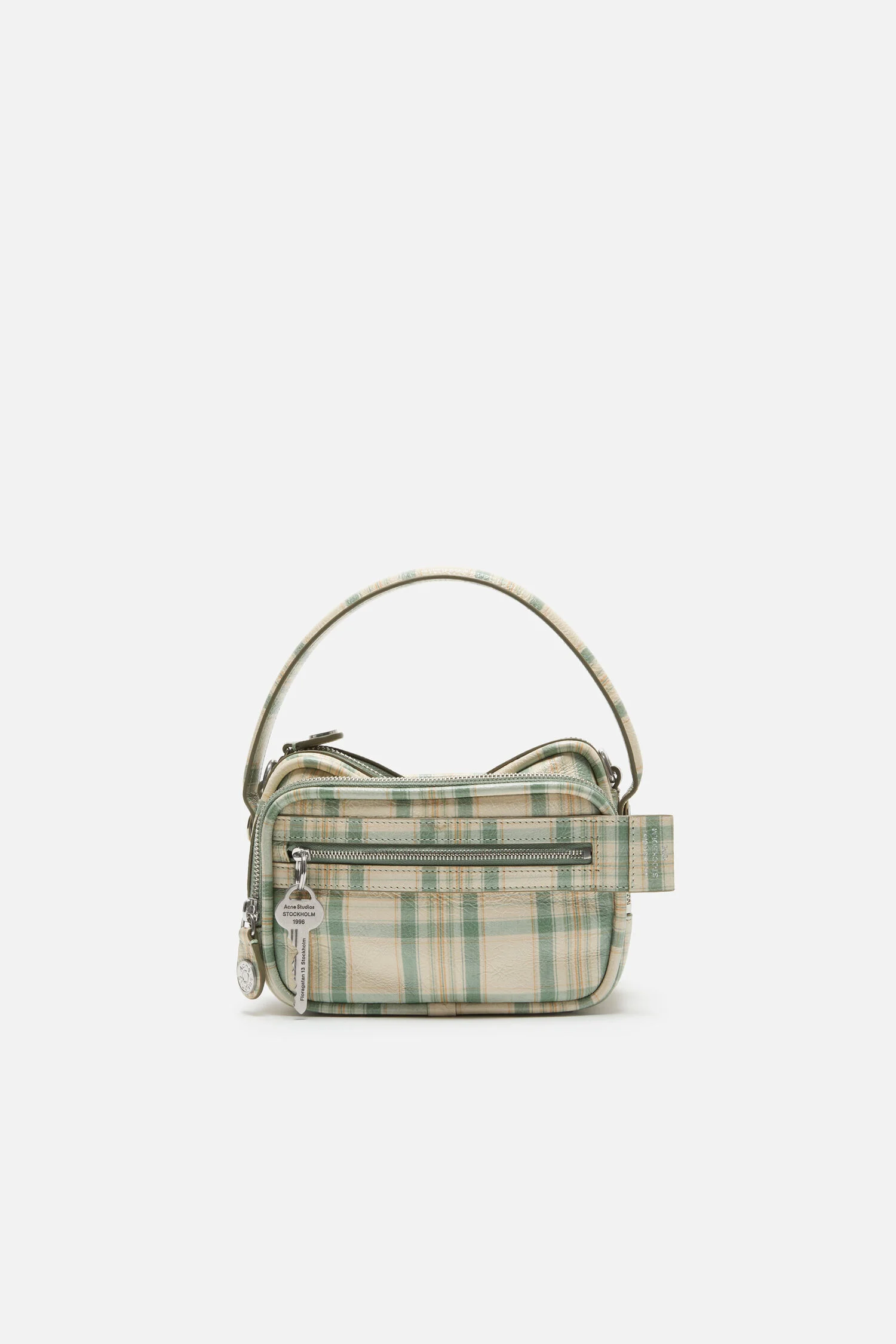 Studio product shot of a compact handbag against a clean light grey background. The bag features a soft rectangular silhouette in a muted green and beige plaid pattern, with a structured top handle and silver-tone hardware. A front zip pocket adds function, accented by dangling branded key tags and zipper pulls. The design blends a classic tartan motif with a modern, minimal shape, emphasizing texture, detail, and everyday practicality