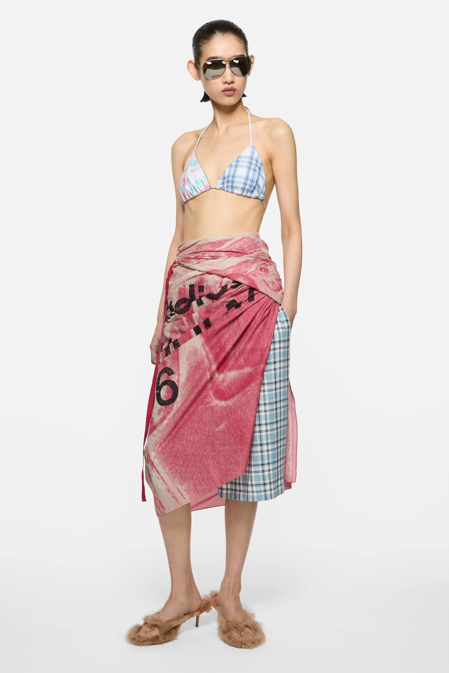 Full-length studio portrait of a model against a clean light grey background, styled in a layered, deconstructed summer look. She wears a delicate triangle bikini top in mixed pastel plaid and soft gradient tones, paired with a wrap-style skirt that combines a bold red printed fabric with visible typography and a contrasting blue plaid panel beneath. The skirt drapes asymmetrically, creating movement and a patchwork effect. Accessories include reflective aviator sunglasses, adding a sharp, modern edge. On her feet are tan, fur-trimmed heeled sandals, introducing texture and softness. The overall composition balances minimal styling with experimental layering, highlighting contrasts in print, fabric, and silhouette