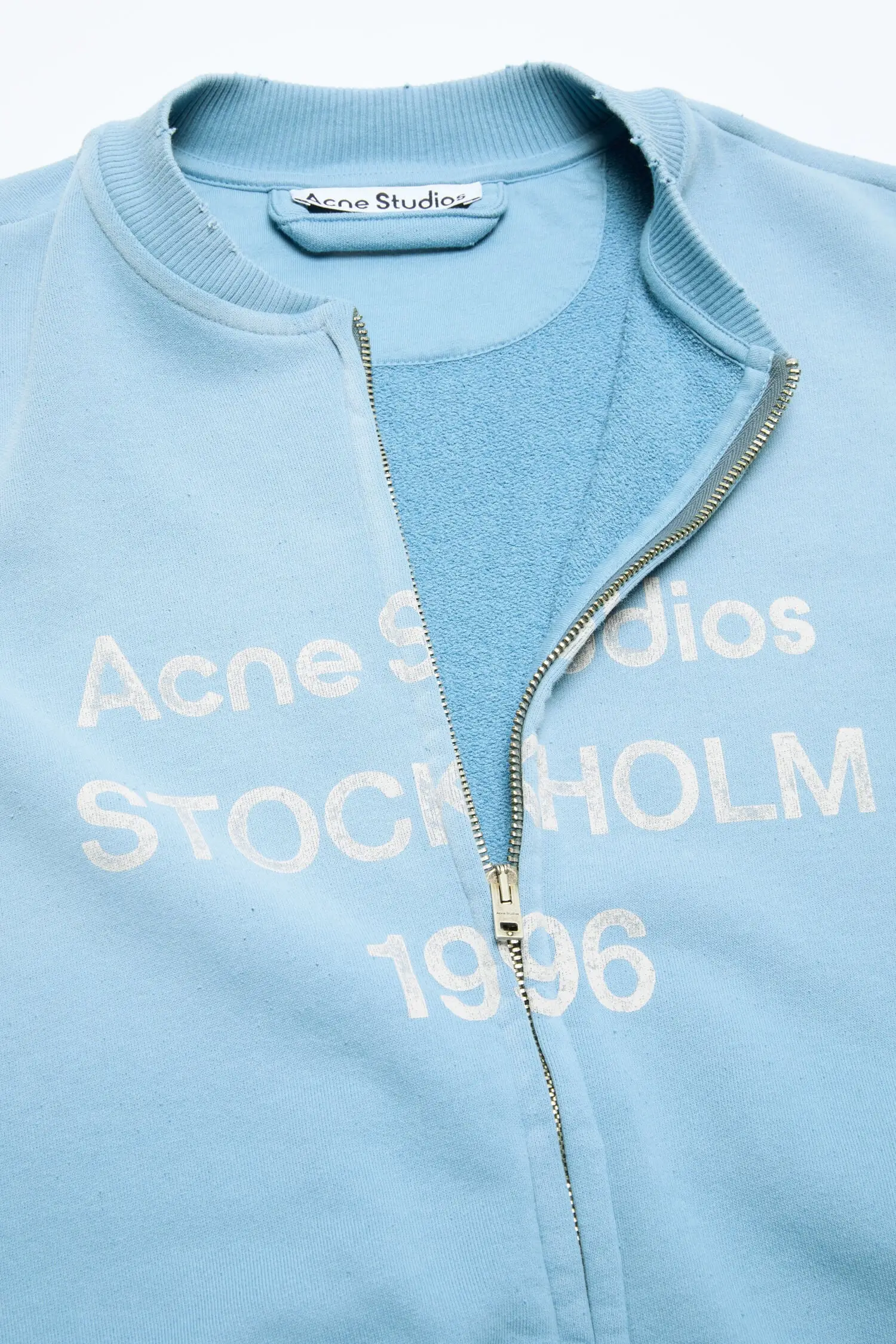 Close-up studio shot of a light blue zip-up sweatshirt against a clean neutral background. The design features a soft fleece interior, ribbed collar, and a partially unzipped silver-tone front closure. White printed text across the chest reads “Acne Studios Stockholm 1996,” with the brand label visible at the neckline. Subtle distressing around the collar edge adds a worn-in detail, balancing the clean color and minimal construction