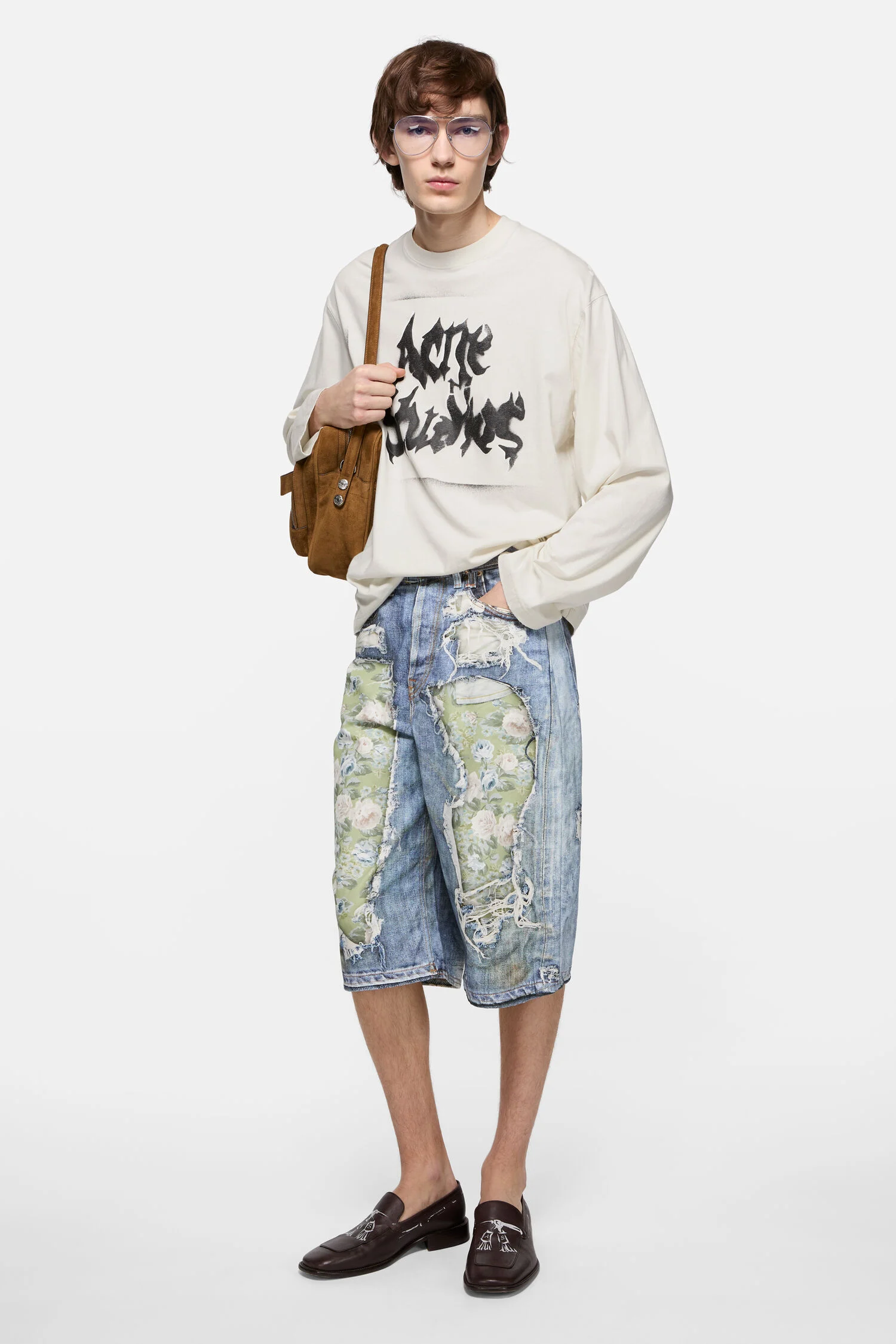 Full-length studio portrait of a model against a clean light grey background, styled in a relaxed, deconstructed casual look. He wears an oversized cream long-sleeve T-shirt featuring a bold black graphic text print across the chest. The top is paired with knee-length denim shorts heavily distressed and patched with green floral fabric, creating a contrast between rugged wear and soft patterning. Accessories include clear-framed eyeglasses and a brown suede shoulder bag worn across one side. On his feet are dark brown leather loafers, adding a polished, slightly preppy finish to the otherwise undone aesthetic