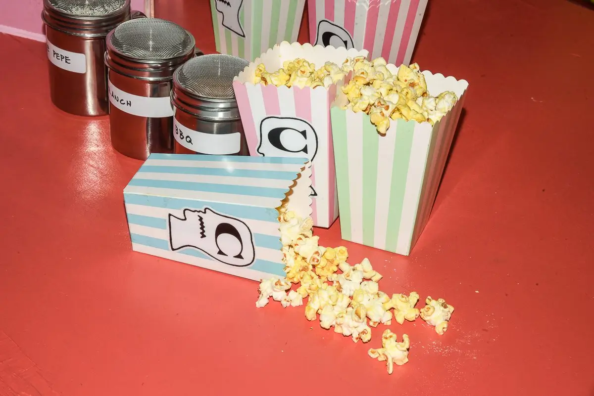 Close-up event detail featuring pastel-striped popcorn boxes decorated with playful graphic illustrations and bold letter motifs. Several containers overflow with buttered popcorn, with scattered pieces spread across a vivid coral floor. Nearby metal seasoning shakers labeled with flavors add to the immersive, carnival-inspired atmosphere, reinforcing the collection’s nostalgic amusement-park and collectible culture aesthetic
