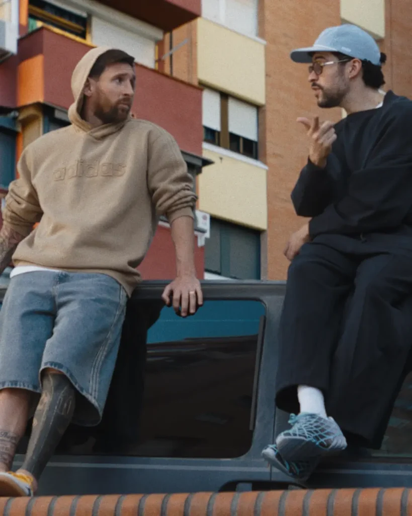 Two men sit casually on top of a parked vehicle in an urban residential setting, engaged in conversation against the backdrop of colorful apartment buildings. One wears a beige adidas hoodie with oversized denim shorts and visible leg tattoos, while the other is dressed in loose black layered streetwear paired with a pale cap, tinted glasses, and futuristic adidas sneakers accented in icy blue. The relaxed body language and muted cinematic color grading create a grounded neighborhood atmosphere that blends football culture, contemporary street fashion, and day city life