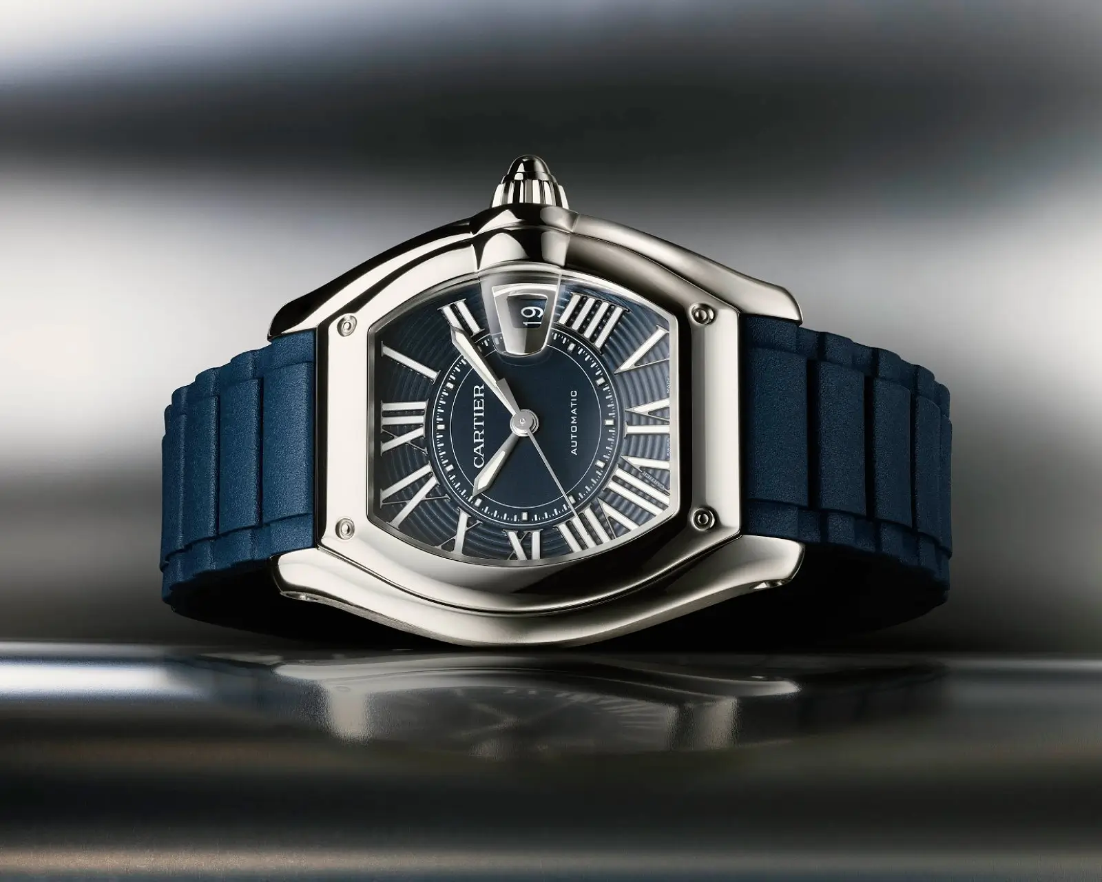 Studio close-up of the Cartier Roadster 2026 in stainless steel with a deep navy blue dial and matching textured rubber strap. The watch showcases the collection’s curved tonneau-shaped case, oversized fluted crown, elongated Roman numerals, integrated magnified date display, and automotive-inspired instrument-panel design language against a sleek metallic background
