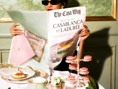 A stylish guest in dark sunglasses reads a custom Casablanca x Ladurée newspaper while seated at a lavish Parisian café table surrounded by signature macarons and pastry