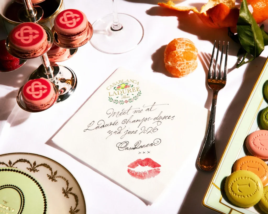 A handwritten invitation from the Casablanca x Ladurée collaboration rests among branded macarons, citrus fruit, elegant silverware, and haute pâtisserie presentation