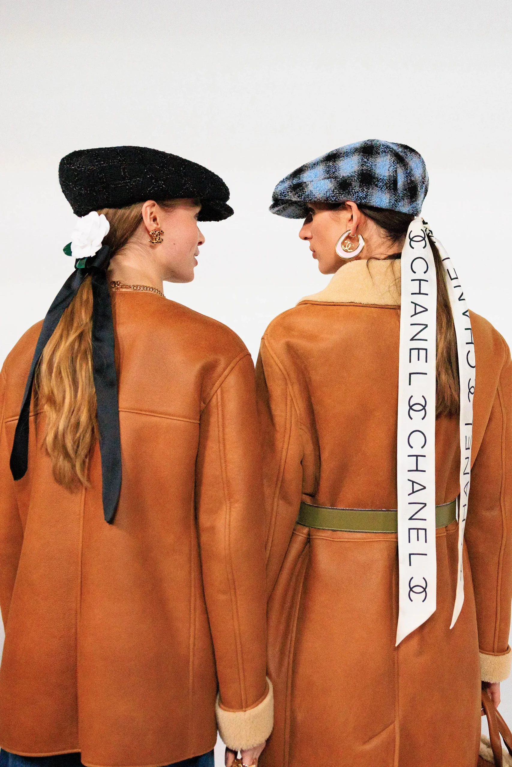 Two models are photographed from behind wearing coordinated Chanel outerwear in warm camel tones with shearling-trimmed finishes. The styling is accented with textured newsboy caps, oversized gold jewelry, and elongated ribbon hair accessories—one featuring oversized Chanel typography—creating a refined mix of equestrian influence, Parisian heritage, and contemporary house styling
