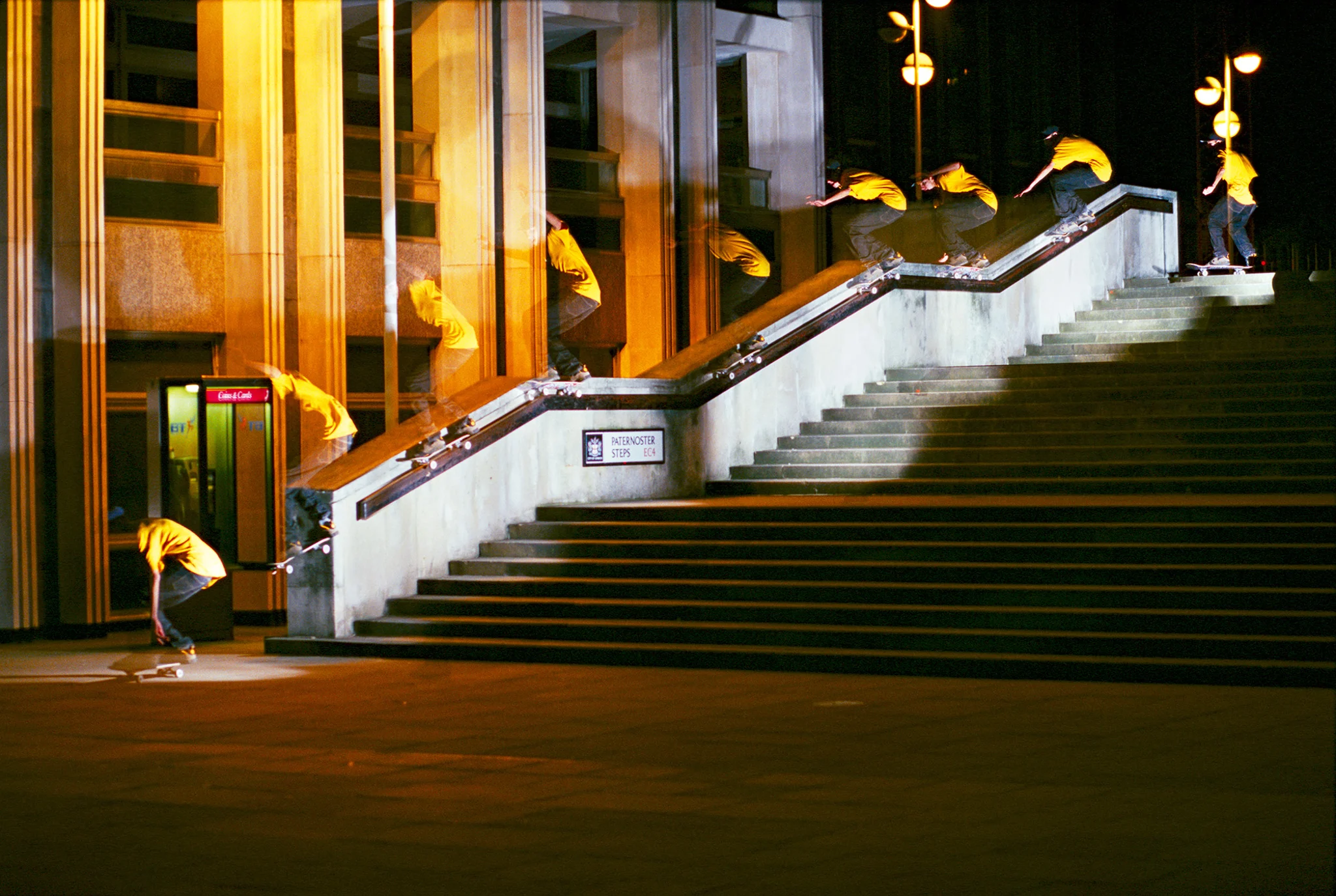 Long-exposure archival skateboarding photograph showing a skater descending a large urban stair set at night, captured in multiple sequential positions along the handrail to emphasize motion, speed, and technical precision against illuminated city architecture and dramatic silhouette