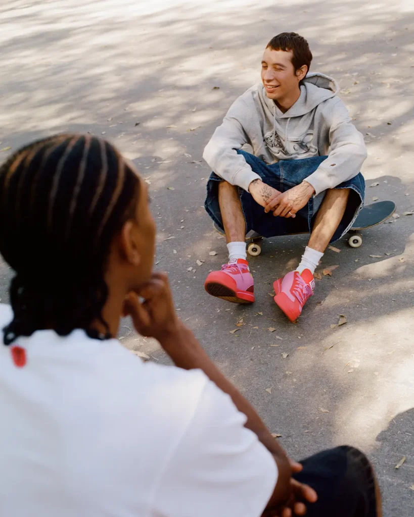 Two friends relax at a sunlit skate spot, captured in a candid moment of conversation and downtime. One skater sits low on a skateboard wearing an oversized gray hoodie, long denim shorts, white socks, and bright pink sneakers that sharply contrast against the muted concrete surroundings. In the foreground, another figure with braided hair appears softly out of focus, adding depth and an intimate documentary feel to the composition. Dappled shadows from nearby trees scatter across the pavement, reinforcing the laid-back, early-2000s-inspired skate aesthetic