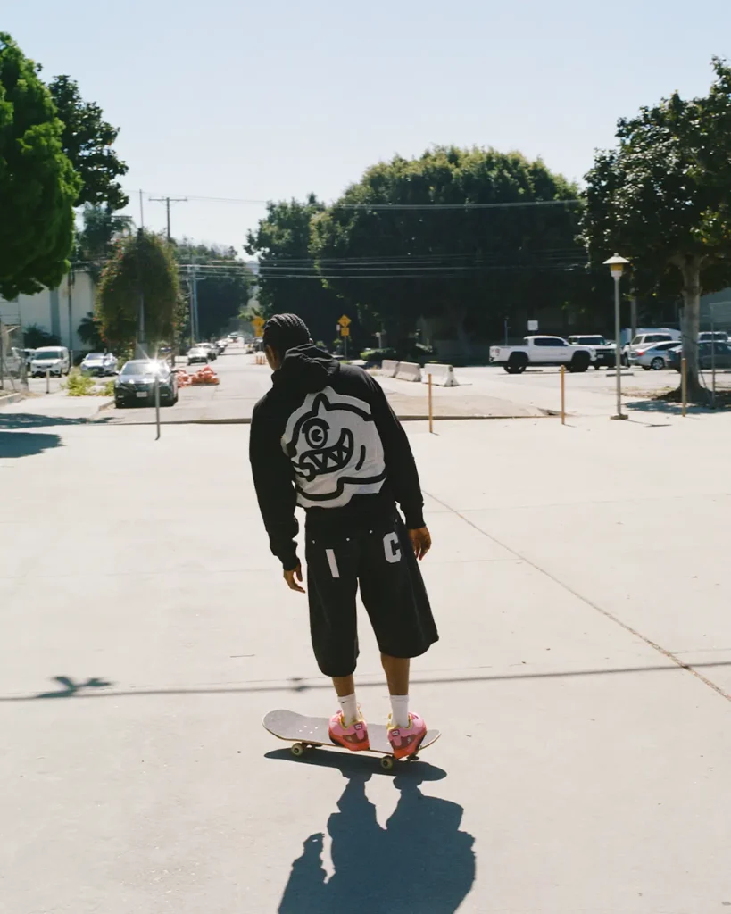 A skateboarder rides through a quiet sunlit street wearing an oversized black hoodie with a bold cartoon-style graphic printed across the back. Loose black shorts, white crew socks, and vivid pink sneakers amplify the look’s playful skatewear energy, while long afternoon shadows stretch across the pavement beneath the board. Captured from behind, the image carries a relaxed documentary feel, blending suburban calm with expressive street-style attitude and early-2000s-inspired skate culture aesthetics
