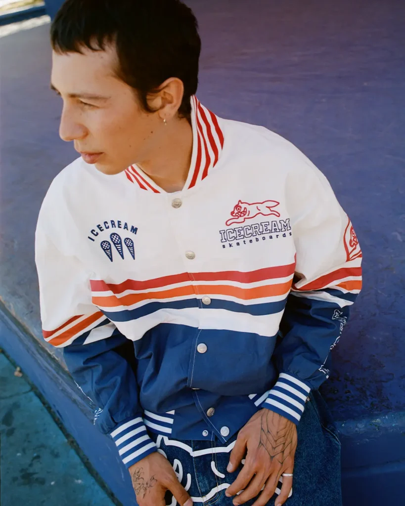 A model sits against a deep blue skatepark ramp wearing a retro-inspired varsity jacket from ICECREAM Skateboards. The jacket combines white, red, and navy color blocking with striped ribbed trims and embroidered graphics across the chest and sleeves, channeling vintage motorsport and collegiate influences through a skatewear lens. Loose denim, visible hand tattoos, and a softly sun-faded color palette give the portrait an effortless early-2000s energy, while the candid side profile reinforces the laid-back authenticity of contemporary skate culture styling