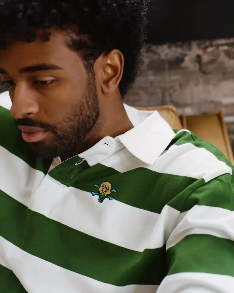 A close-up portrait captures a model wearing a green-and-white striped rugby polo with a small embroidered cartoon graphic positioned at the chest. The heavyweight shirt blends classic collegiate sportswear references with playful streetwear detailing, while the crisp white collar adds structure against the relaxed silhouette. Soft indoor lighting and the textured industrial backdrop create a warm, intimate atmosphere, emphasizing the rich fabric texture and understated vintage-inspired styling