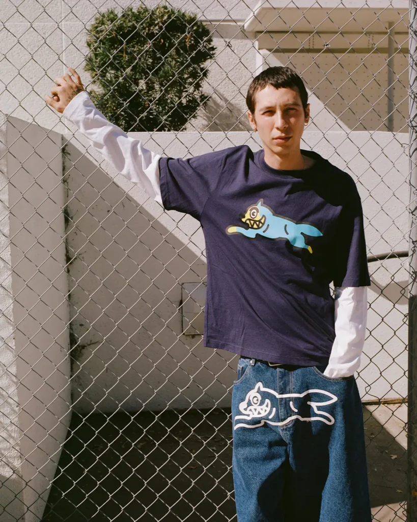 A model poses casually beside a chain-link fence wearing layered skate-inspired streetwear under bright afternoon sunlight. The outfit centers around a navy graphic T-shirt layered over a white long-sleeve shirt, paired with oversized denim shorts featuring a large cartoon dog illustration across the front. The matching graphics create a cohesive visual identity rooted in playful skate culture, while the relaxed proportions, washed denim textures, and stark urban backdrop reinforce a nostalgic early-2000s streetwear mood