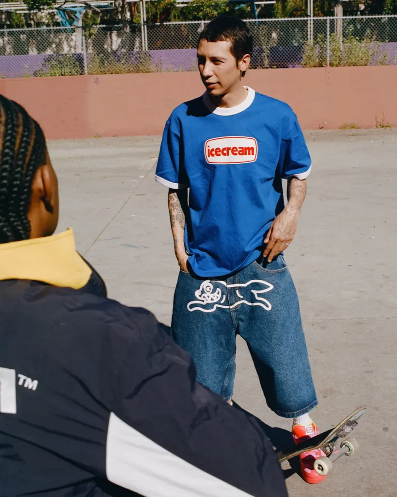A candid skatepark scene captures a model wearing a vibrant blue ringer T-shirt from ICECREAM Skateboards paired with oversized denim shorts featuring the brand’s signature running dog graphic across the front. Bright pink sneakers add a sharp pop of color against the faded concrete setting, while visible tattoos and relaxed body language reinforce the effortless streetwear attitude. Framed alongside another skater in the foreground, the image carries a documentary-style energy rooted in community, movement, and nostalgic skate culture aesthetics