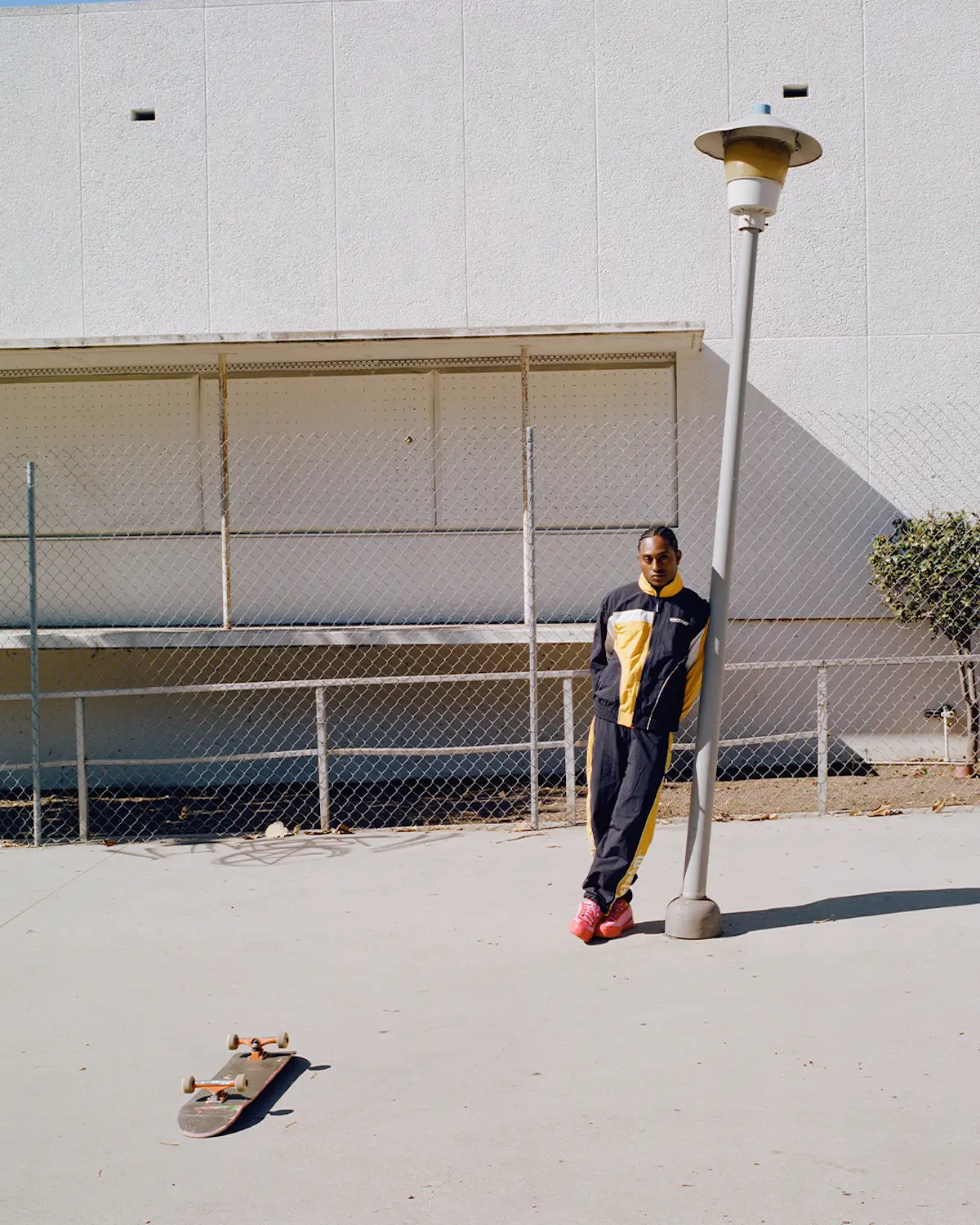 A wide editorial-style shot captures a model leaning against a streetlamp in a coordinated black-and-yellow tracksuit from ICECREAM Skateboards. The sporty set combines retro athletic influences with contemporary skatewear styling, accented by vivid pink sneakers that stand out against the pale concrete surroundings. A skateboard rests in the foreground, reinforcing the relaxed skate-culture atmosphere, while the stark industrial architecture, chain-link fencing, and strong midday sunlight give the composition a cinematic sense of openness and urban minimalism