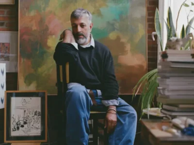 Portrait of photographer Daniel Riera seated inside a warmly lit studio-like interior surrounded by stacked books, framed artworks, plants, and expressive paintings. Wearing relaxed blue trousers, a dark knit sweater, and woven leather sandals, Riera appears contemplative against a richly textured artistic backdrop, capturing the intimate atmosphere of a creative life shaped by collecting, design, and Mediterranean culture sensibility