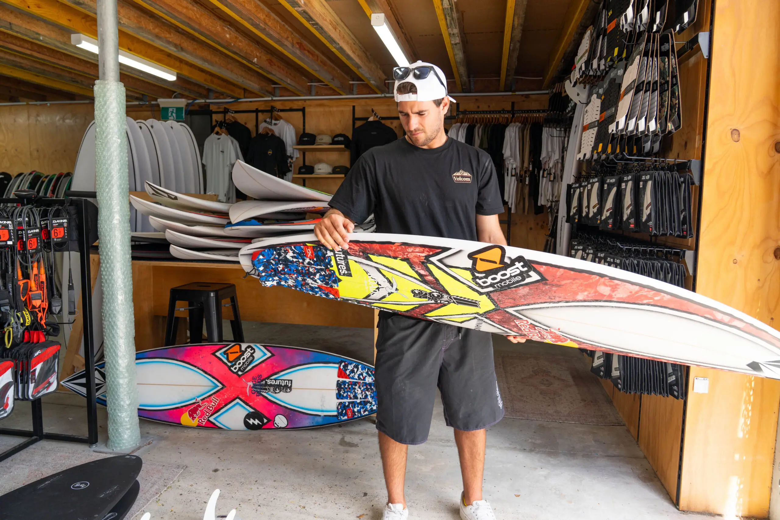 A lifestyle-driven surf retail image featuring a rider inside a wood-paneled board shop carefully inspecting a high-performance shortboard covered in sponsor decals and vibrant traction detailing. Surrounded by stacked surfboards, wetsuit accessories, and apparel racks, the scene captures the culture of surf craftsmanship, equipment selection, and the everyday ritual behind modern wave perform