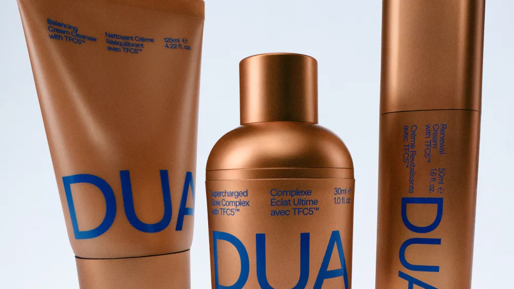 Close-up product lineup of the DUA by AB skincare collection featuring three metallic bronze containers with bold cobalt blue typography. The minimalist packaging highlights the cleanser, glow serum, and renewal cream, emphasizing a modern blend of luxury design and science-led skincare