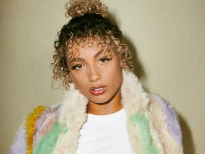 Portrait of singer DaniLeigh wearing a pastel multicolored faux-fur jacket over a white top. She poses against a neutral backdrop with her curly hair styled in a high bun, looking directly at the camera with a confident expression