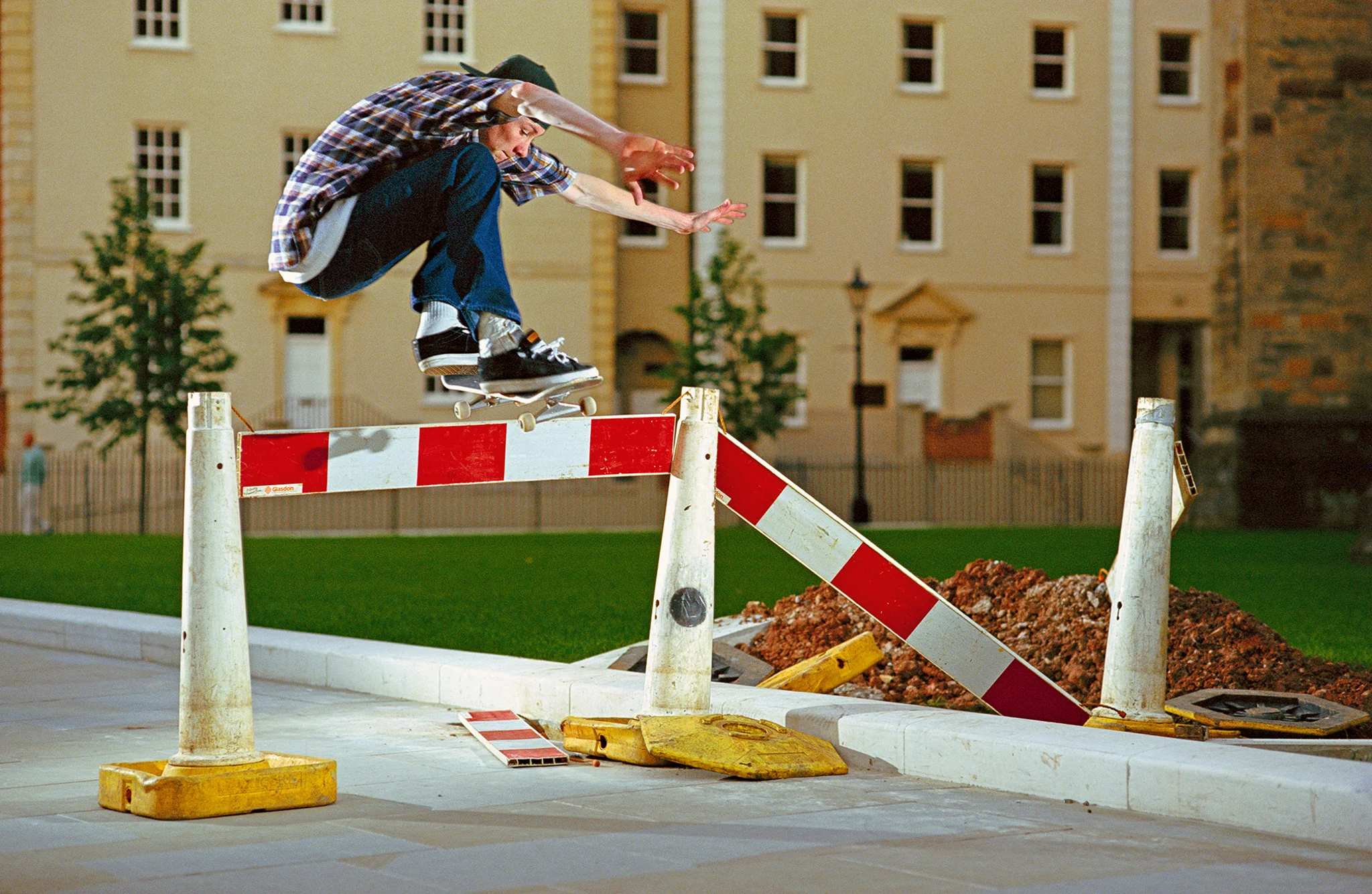 Archival skateboarding photograph capturing a skater clearing a red-and-white construction barrier with a high aerial trick in an urban plaza, framed by historic architecture and open public space—reflecting the inventive, obstacle-driven character of classic UK street skating