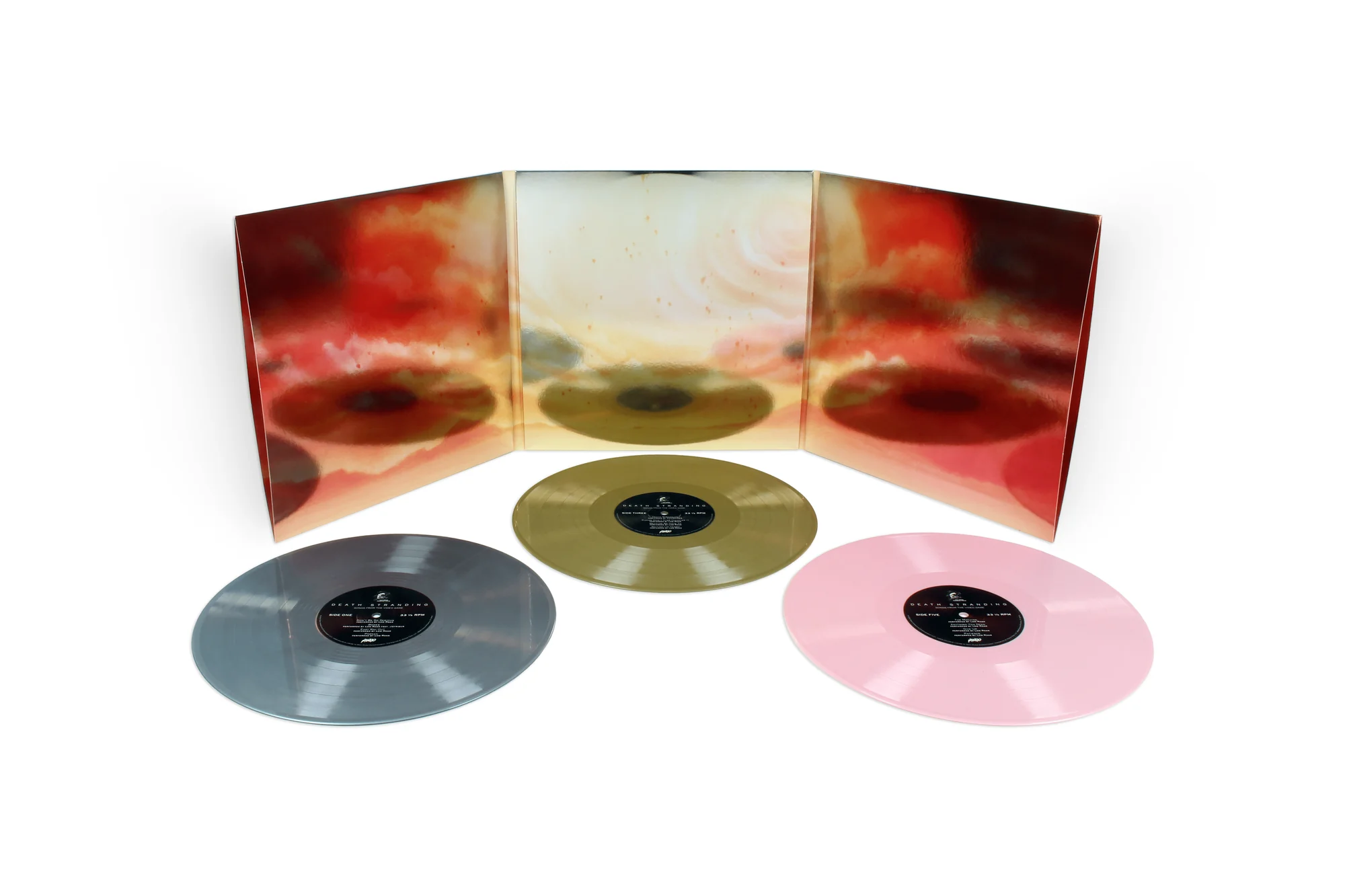Collector’s edition Death Stranding soundtrack vinyl set displayed against a clean white background, featuring a panoramic tri-fold gatefold sleeve with abstract red, orange, cream, and smoky cloud artwork. Three colored records are arranged in front: a translucent silver-gray LP, a translucent gold LP at center, and a soft translucent pink LP, each with minimalist black center labels that reflect the atmospheric, cinematic tone of the game’s soundtrack