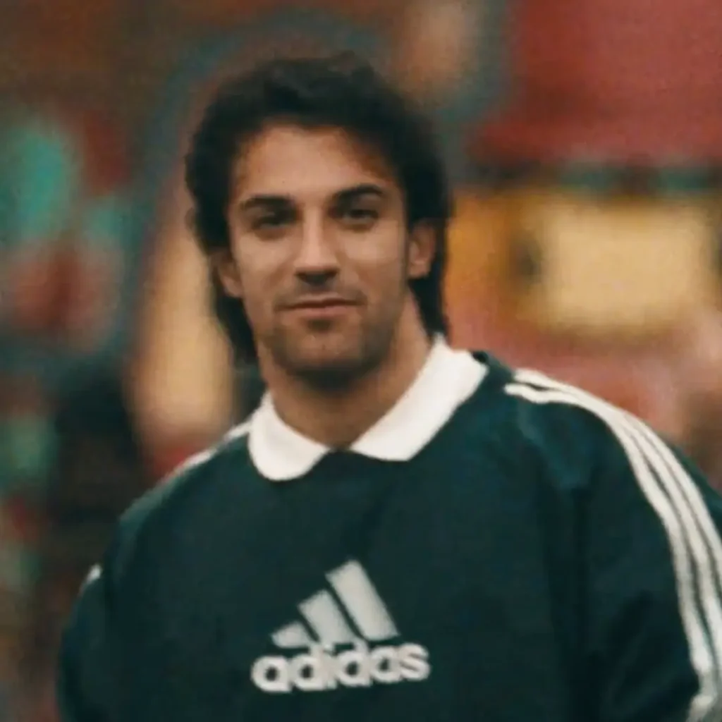 A young man is captured in a softly blurred, vintage-style portrait wearing a dark green adidas football sweatshirt with classic white three-stripe detailing along the sleeves and a white polo collar underneath. The close-up composition and warm grainy texture give the image a nostalgic late-1990s sportswear aesthetic, while the muted urban background fades into soft color washes behind him. His relaxed expression and timeless football styling evoke the atmosphere of classic European street football culture and archival adidas campaign imagery
