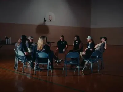 Members of Dexter and The Moonrocks and Treaty Oak Revival sit in a circle of folding chairs inside a dimly lit gymnasium, evoking the visual language of a support group meeting that mirrors the recovery themes explored in their collaborative single “12 Steps.”