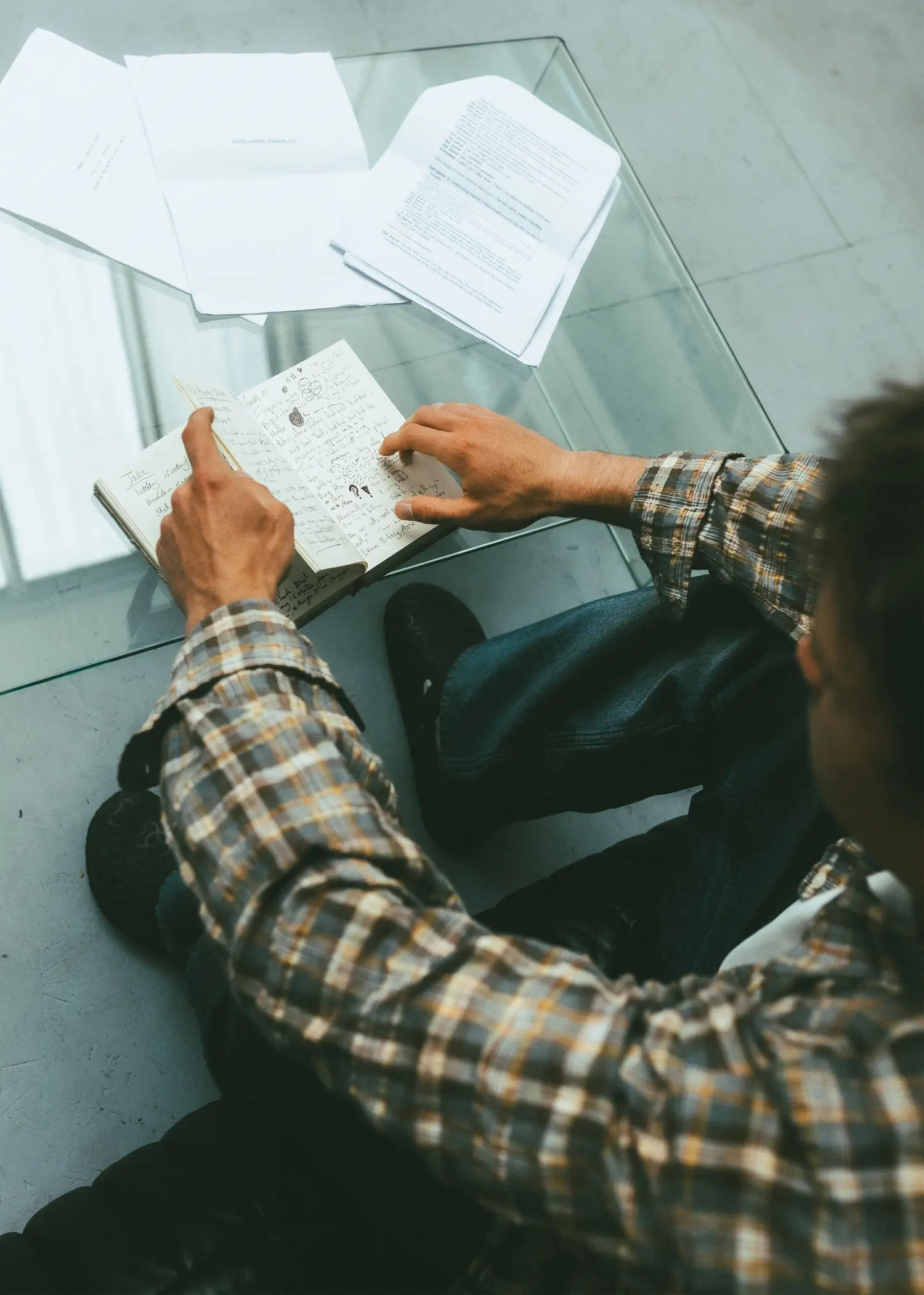 Overhead lifestyle image of a person seated at a glass table reviewing handwritten notes in an open notebook, surrounded by printed documents and papers. The relaxed setting, layered plaid shirt, and editorial workspace atmosphere emphasize process, reflection, and creative development in a quiet studio-like environment