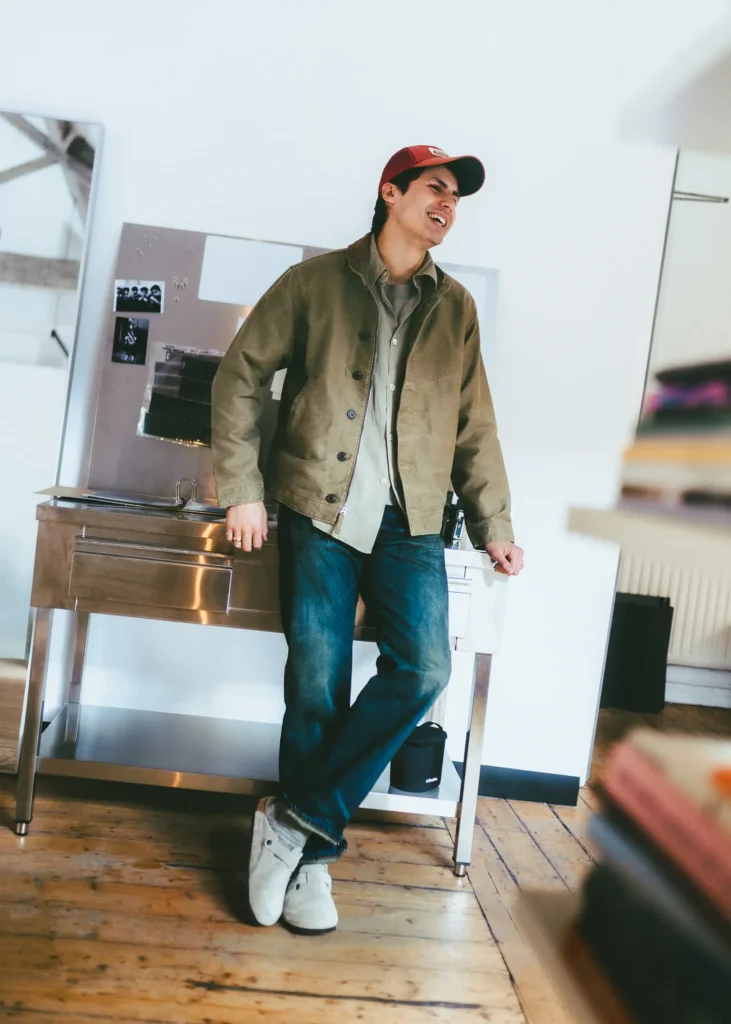 Lifestyle image of a person leaning casually against a stainless steel utility table in a bright loft-like interior, wearing light beige suede clogs with socks, dark denim, layered neutral outerwear, and a red cap. Warm wooden flooring, soft daylight, and relaxed styling create an effortless everyday aesthetic centered on comfort and understated design